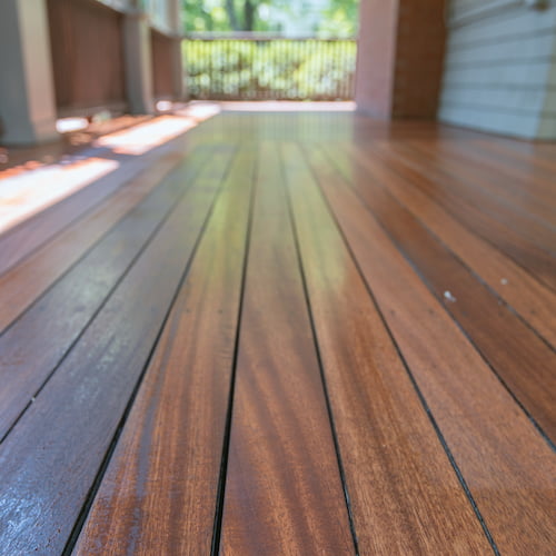 Close-up perspective view of a polished wooden floors with rich brown tones, leading towards a blurred background of greenery and a porch railing.