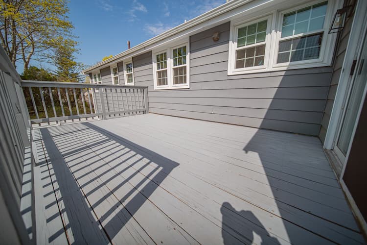 Outdoor hardwood floor of a house painted in light gray, featuring a railing with shadows cast by the sun, adjacent to a house with gray siding and white-framed windows.