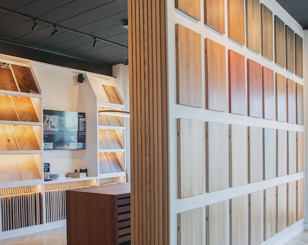 Interior corner of a flooring showroom featuring a wall with various wooden floor samples and a digital display screen.